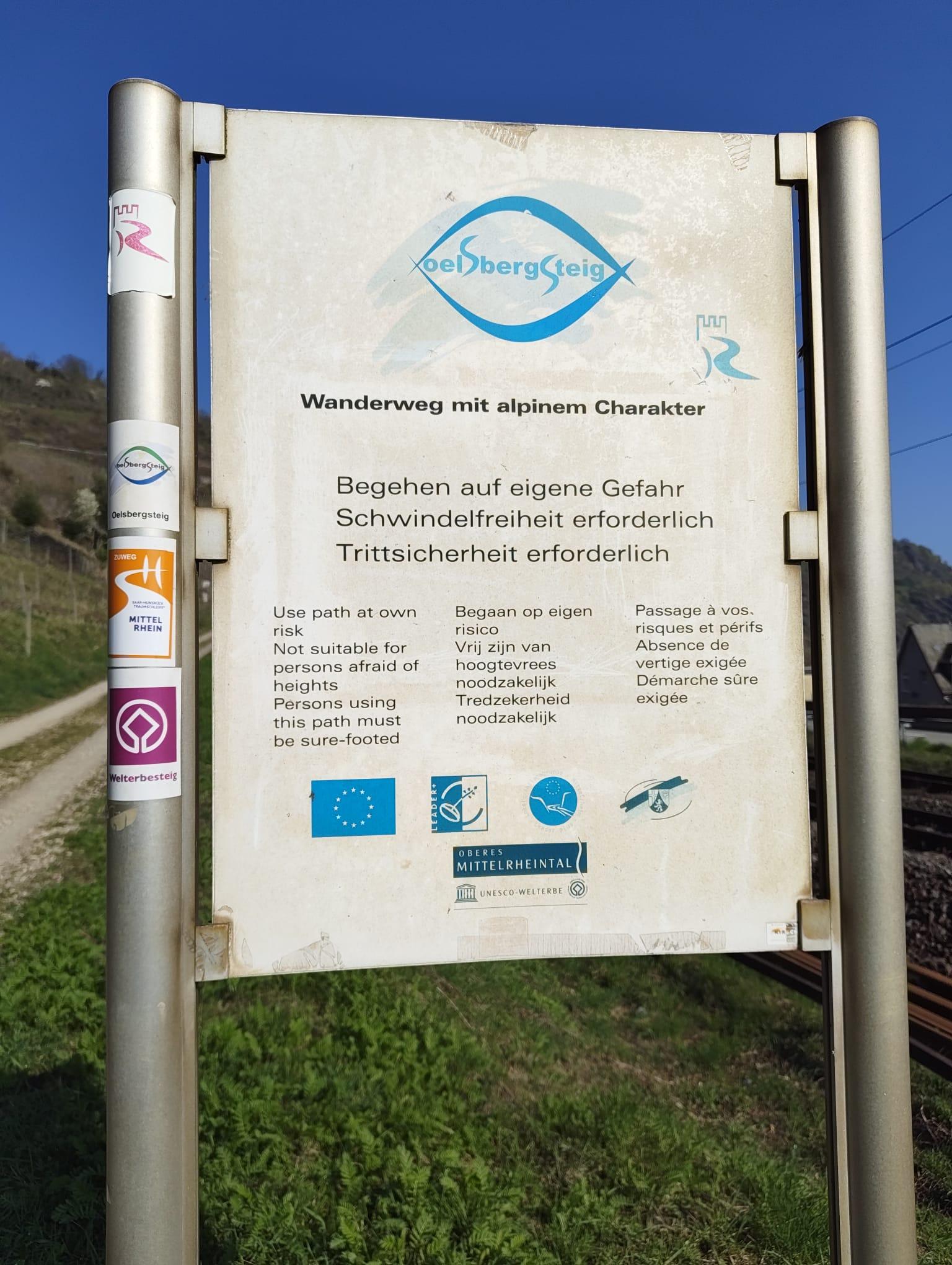 Ein Schild mit der Aufschrift "Wanderweg mit alpinen Charakter,, begehen auf eigene Gefahr, Schwindelfreiheit erforderlich, Trittsicherheit erforderlich". Die Aufschrift findet sich dort auch in anderen Sprachen wieder. Am Schild angebracht sind verschiedene Aufkleber, die die Wanderwege kennzeichnen. Rechts vom Schild befindet sich eine Böschung, neben der eine Bahnlinie verläuft. Rechts vom Schild befindet sich ein Feldweg.