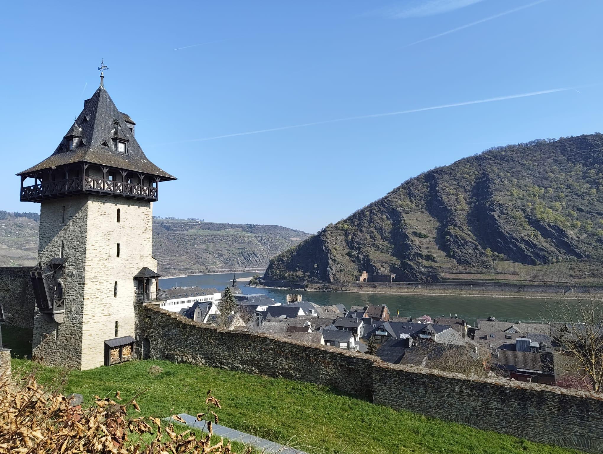 Im vorderen Teil des Bildes befindet sich eine kleine, leicht abschüssige Wiese. Hintendran befindet sich eine mittelalterliche Stadtmauer mit angeschlossenen mittelalterlichen Wachturm mit Spitzdach und kleiner Zugbrücke, der anscheinend restauriert wurde und gut in Schuss ist. Hinter der Stadtmauer und den Turm befindet sich ein kleines Städtchen, hinter dem der Rhein verläuft. Hinter dem Rhein verläuft eine Bahntrasse und eine Straße, bevor sich ein steiler Berg dahinter auftürmt.